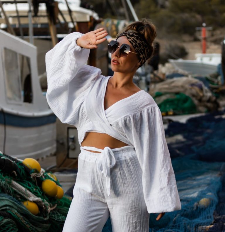 Frau in weißer Kleidung mit Sonnenbrille, die am Hafen im Freien posiert.