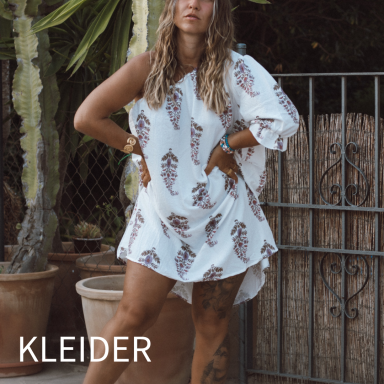 Frau in einem weißen Kleid mit floralem Muster, posiert vor einem Gitterzaun.